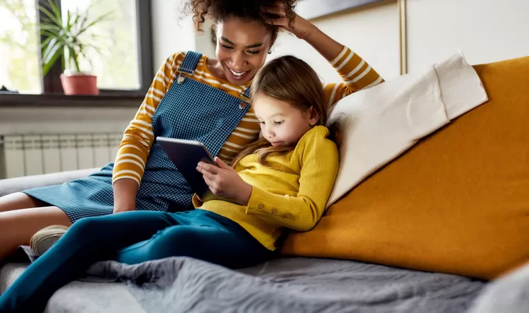 Quality childcare matters. African american woman baby sitter and caucasian cute little girl smiling, using tablet pc, sitting at home. Children education, leisure activities, babysitting concept